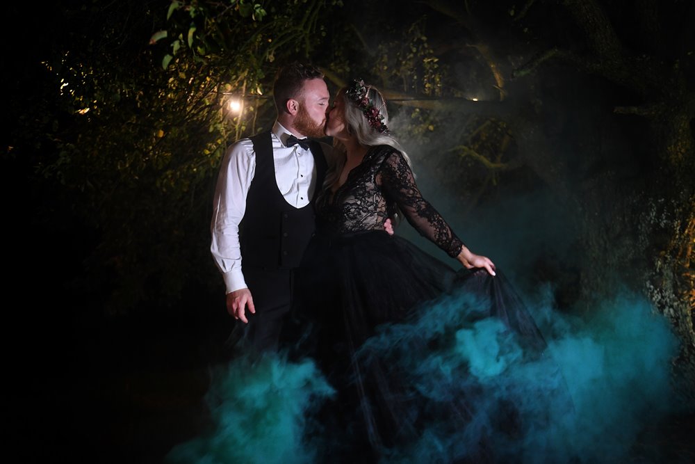 Bride in black wedding dress and autumn flower crown kisses husband in moody photoshoot with smoke bombs at their halloween wedding