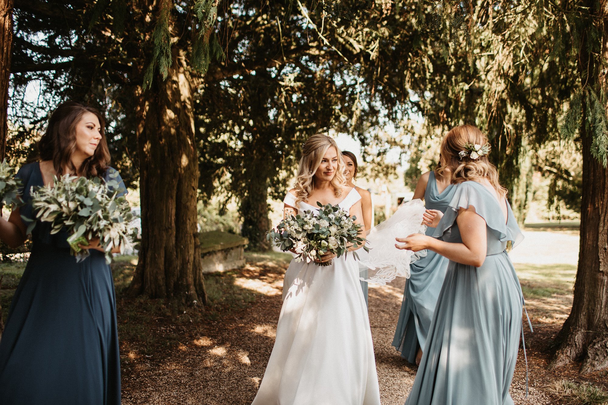 Candid wedding photo of bride with her bridesmaids in teal dresses surrounded by dappled sunshine through the trees