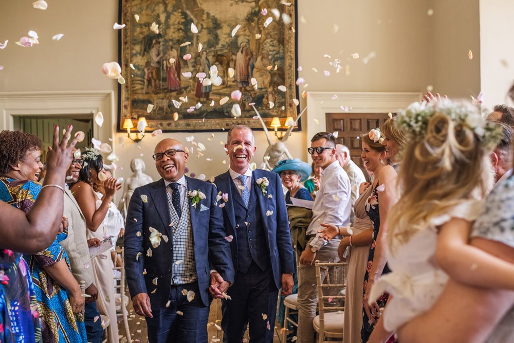confetti thrown as two newlywed grooms walk down the aisle smiling as mr and mr at manor house elmore court