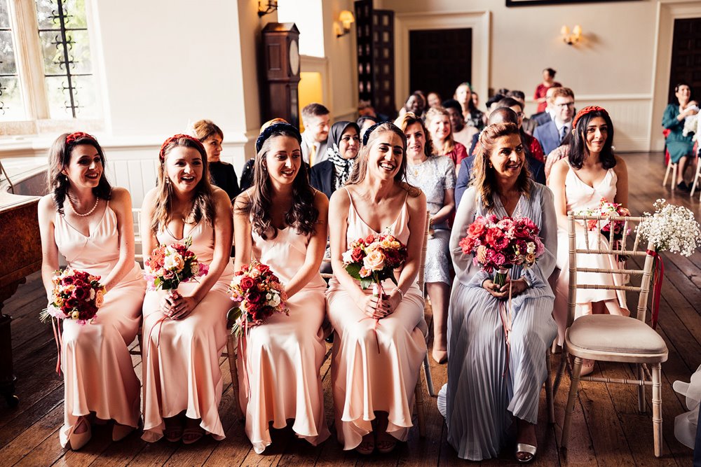 Bridesmaids in pink dresses and red headbands sit in front row of wedding ceremony hall at elmore court
