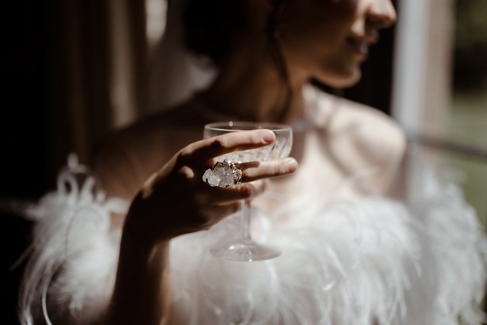 Bride in feathers holding crystal glass with oversized uncut gemstone ring for editorial wedding inspiration