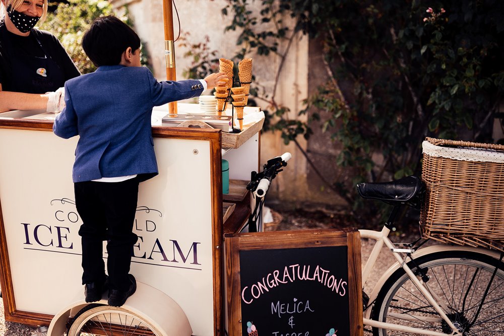 Little boy helps himself to ice cream cone standing on wheel of vintage bicycle ice cream stand at a garden wedding in the cotswolds