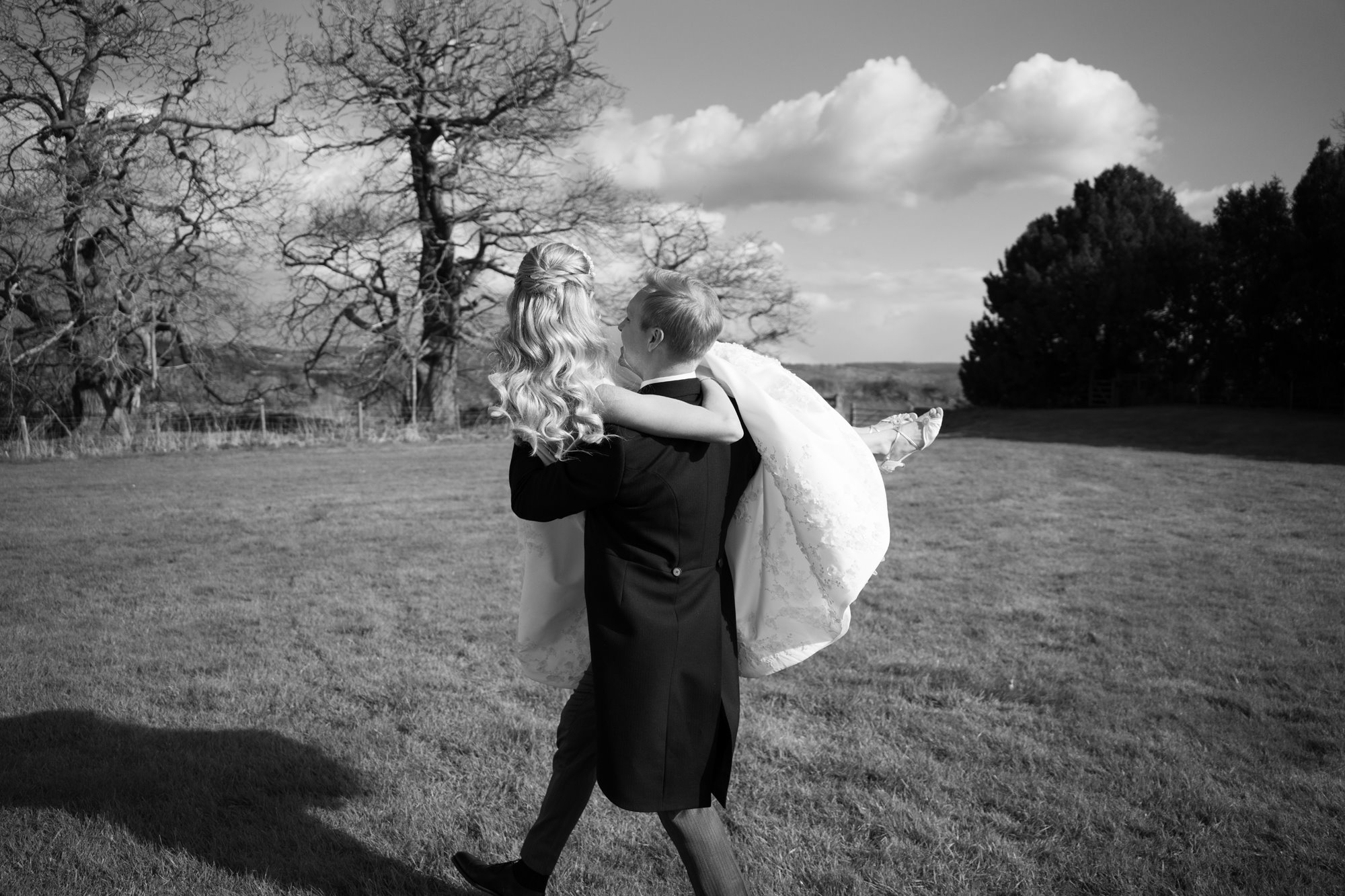 groom carrying his bride across a beautiful countryside lawn with trees in the background