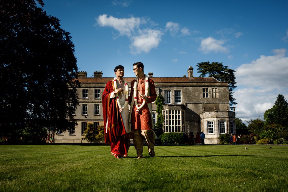 Two grooms in traditional Hindu wedding attire standing in front of mansion house wedding venue Elmore Court at their beautiful gay wedding