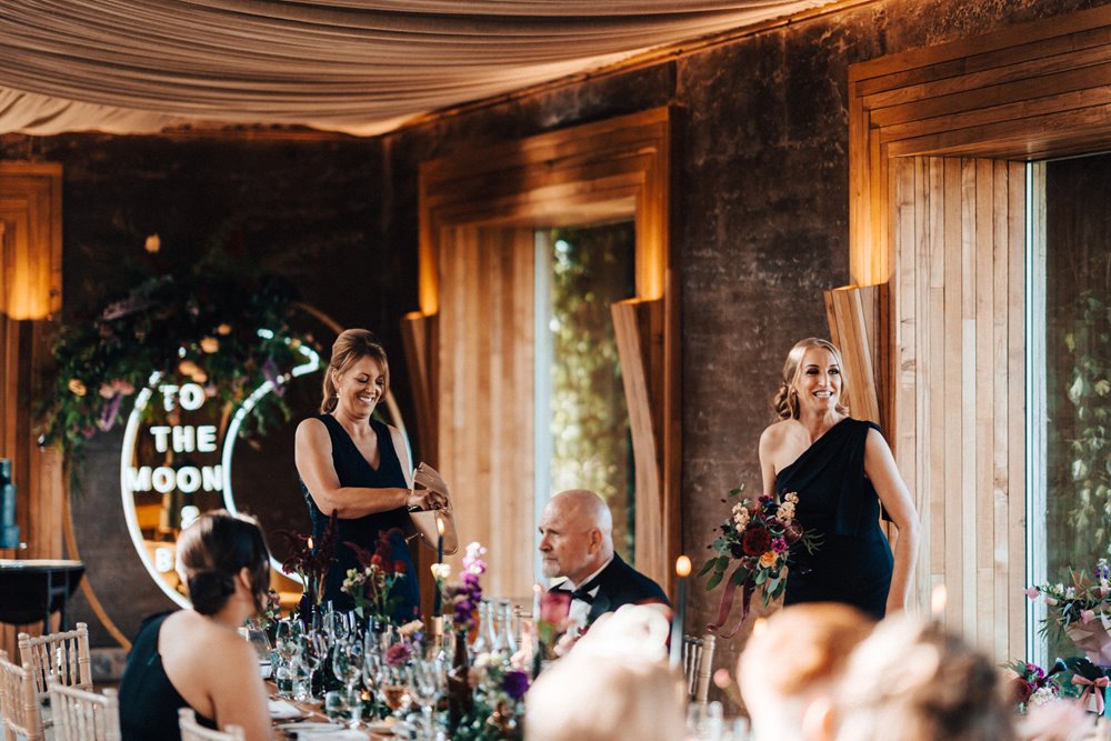 Bridesmaids in black dresses with modern neon to the moon and back sign at an elegant black and gold wedding reception in the cotswolds