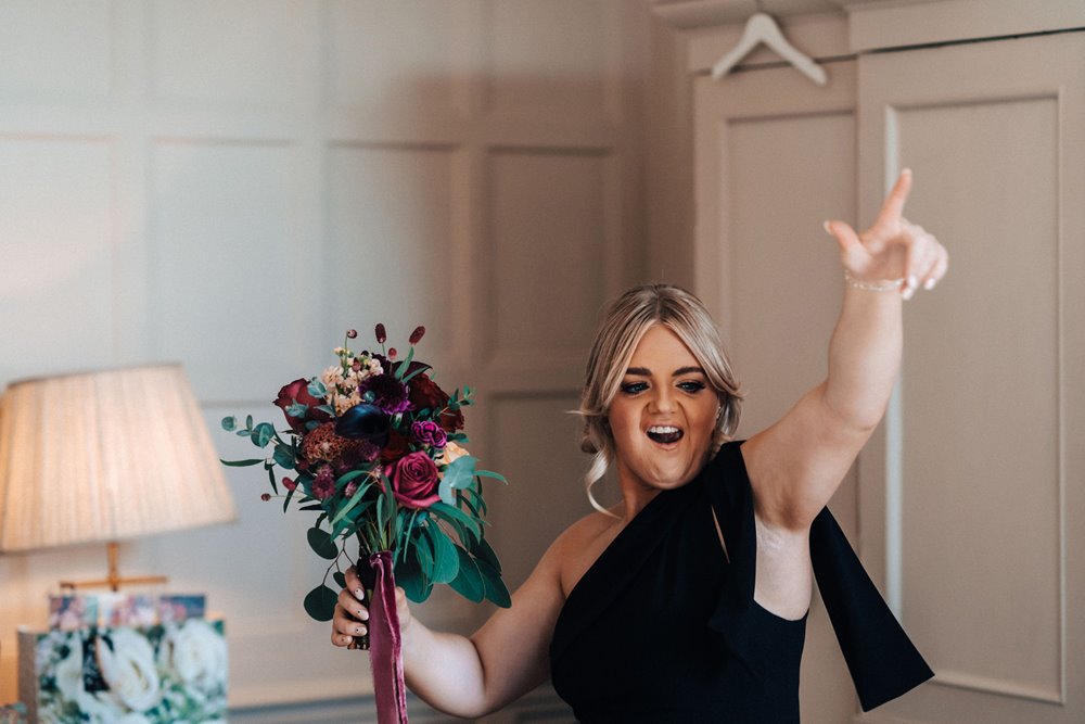 Rock n roll bridesmaid wearing black holds autumn bouquet of dark flowers and dances to music with fingers in the air at mansion party house in England