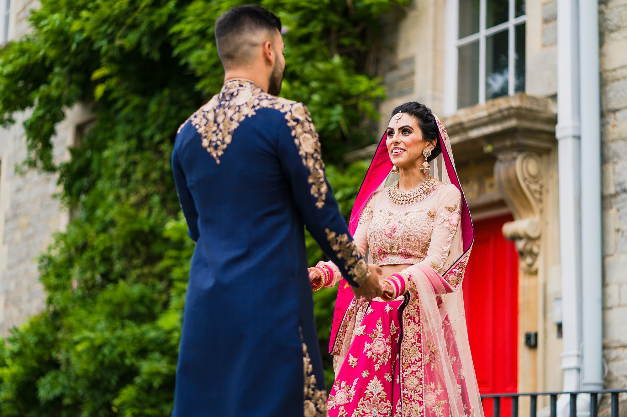 Indian bride and groom dresses in beautiful traditional indian wedding attire take a moment for a first look and gaze lovingly at each other holding hands in front of the red side door at Elmore Court