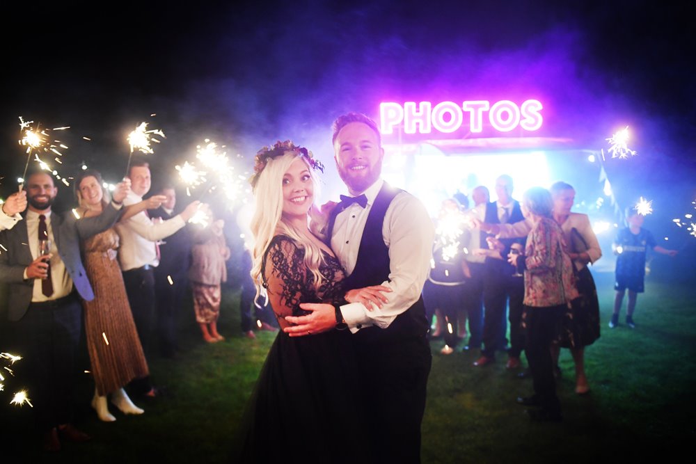 Halloween loving Bride in black wedding dress surrounded by guests with sparklers in front of unusual photo booth at autumn wedding