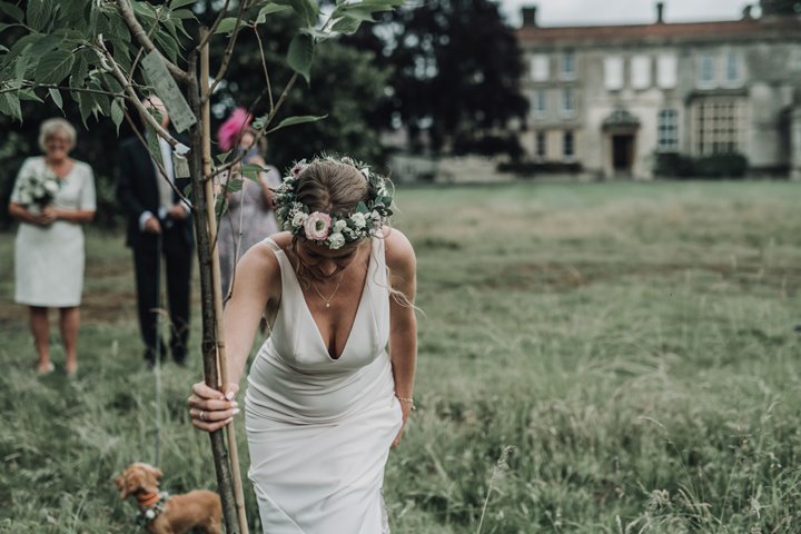 Bride in flower crown and wedding dress leans down to plant her tree with puppy at her feet in her wedding ceremony outside stately home elmore court