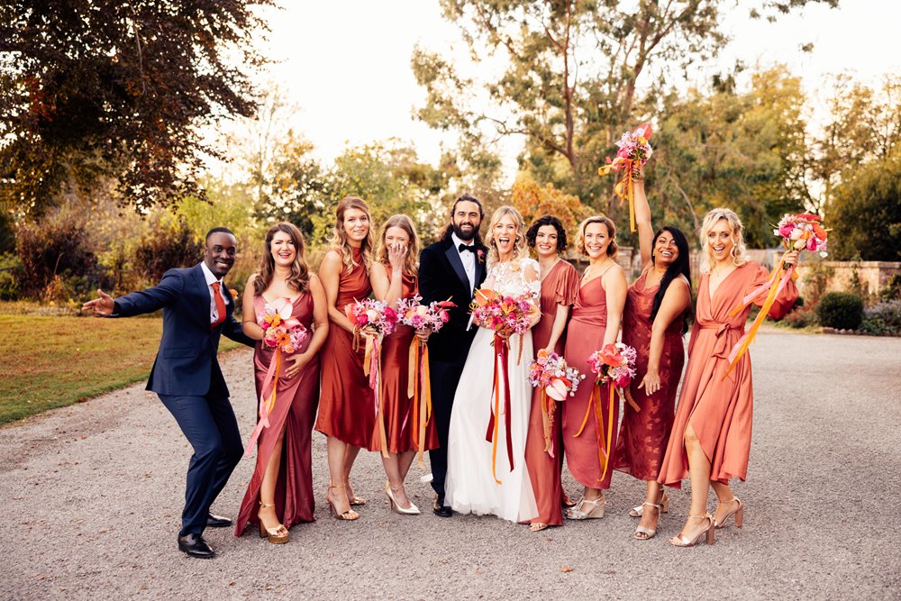 mixed wedding party and bridesmaids in burnt orange copper dresses stand with boho bride with bright bouquets outside stately home wedding venue in the cotswolds countryside 