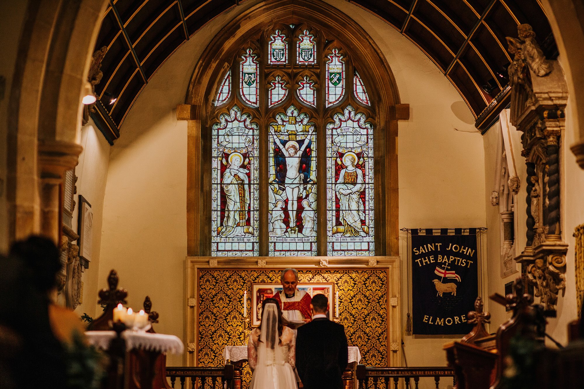 Couple and Vicar in midst of wedding ceremony at the stunning Elmore church in England with high ceilings, stained glass windows and gold details