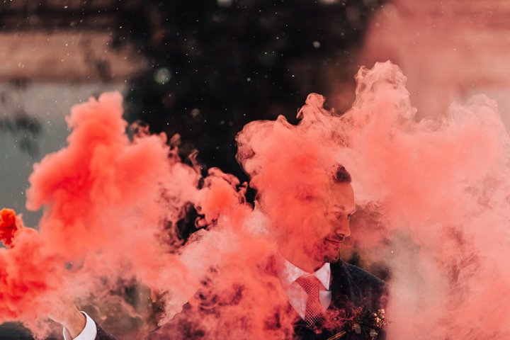 Coral coloured smoke from a smoke bomb obscures part of grooms face in a beautiful cloud of bright colour