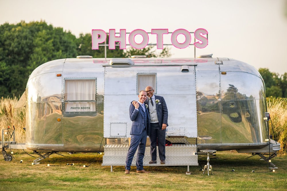 gay couple stand in front of their silverstream caravan photobooth at golden hour in the meadow outside eco wedding venue elmore court