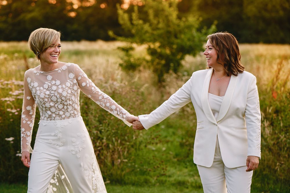 Two brides in white jumpsuit and tuxe hold hands and smile at each other outside in the meadow of perfect same sex wedding venue in Cotswolds countryside