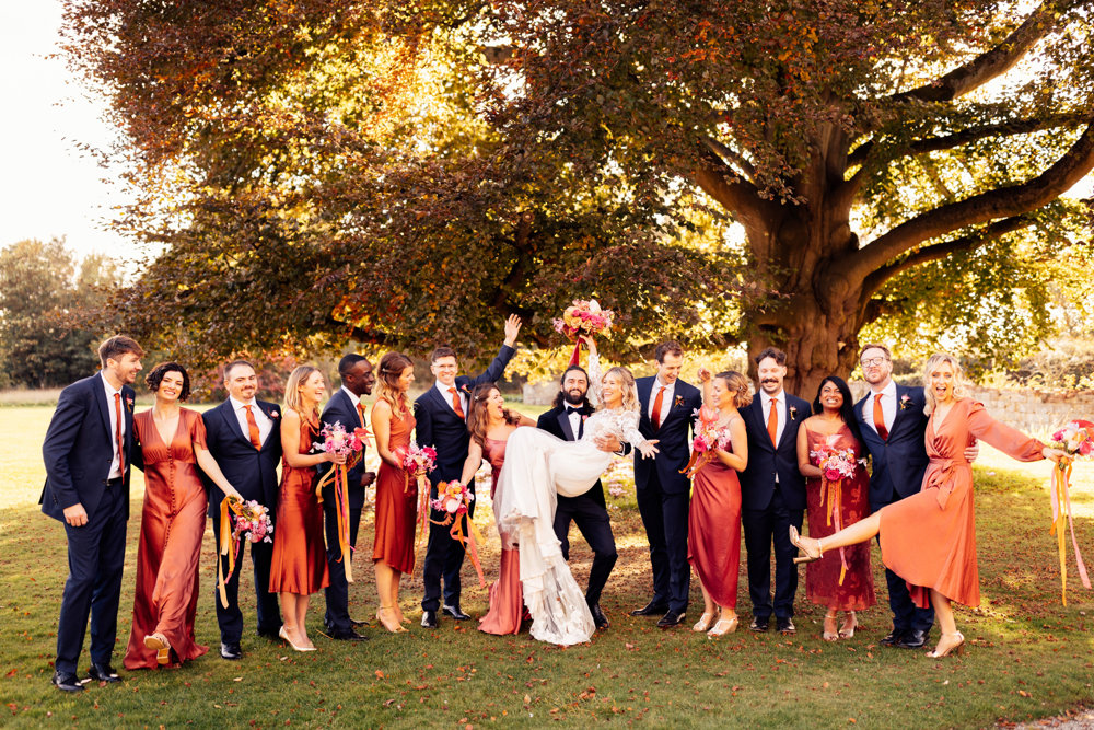 Colourful bridesmaids and groomsmen pose under beech tree with boho bride with bright bouquets with ribbons in the cotswolds countryside