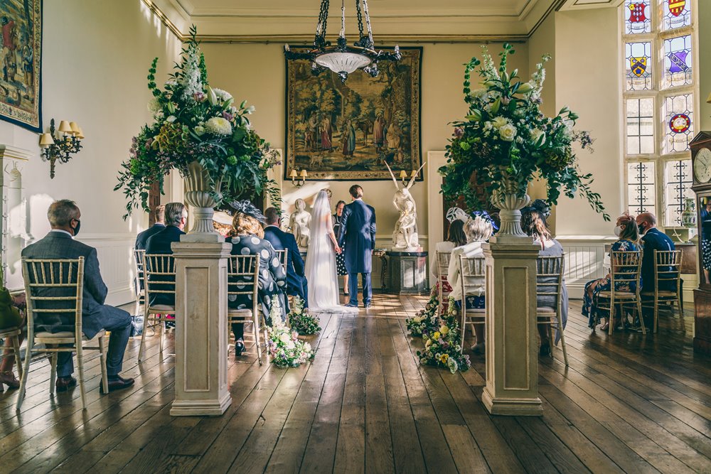 Micro wedding with big styling. Urns full of foliage and flowers flank the wedding aisle in stately home where a small number of guests watch bride and groom marry