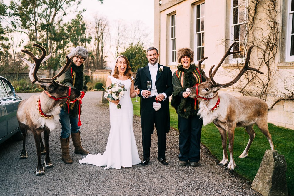 Bride and groom holding champagne pose with reindeer after marrying at Christmas wedding venue elmore court