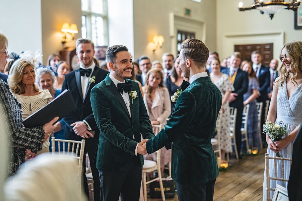 Two grooms in matching green velvet suits holding hands facing each other at the end of the aisle in their gar wedding ceremony in a stately home