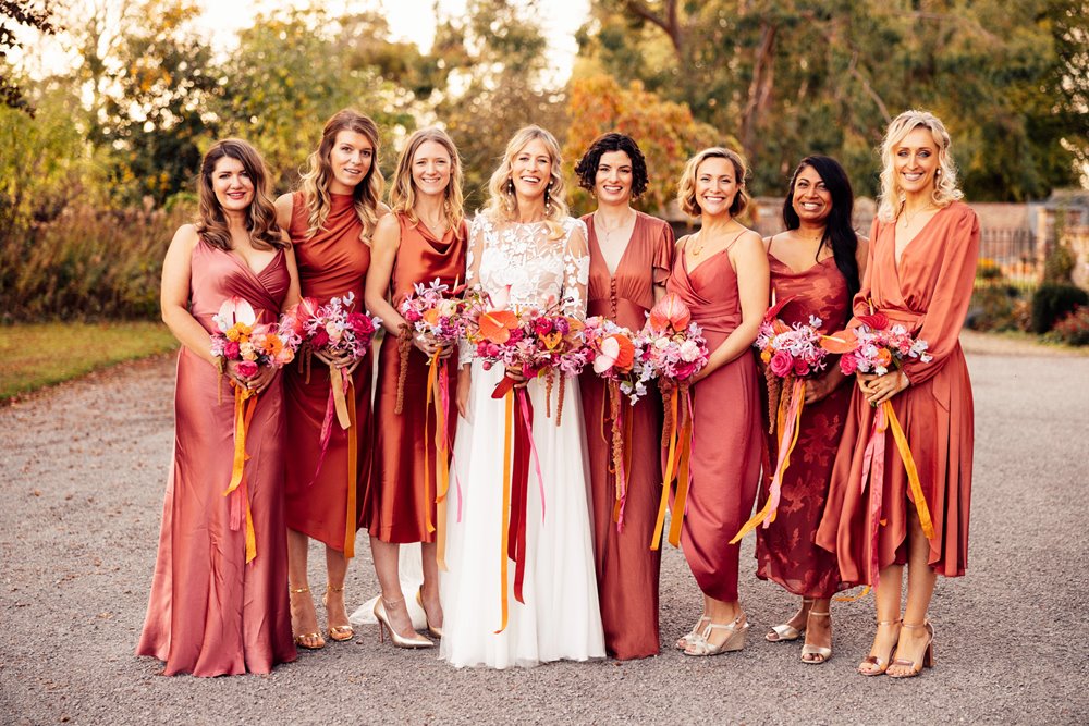 bridesmaids in burnt orange copper dresses stand with boho bride with bright bouquets outside stately home wedding venue in the cotswolds countryside 