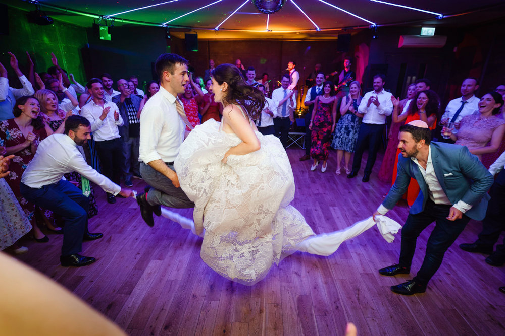 Jewish bride skips in her huge wedding dress holding it bunched up to jump over napkin rope