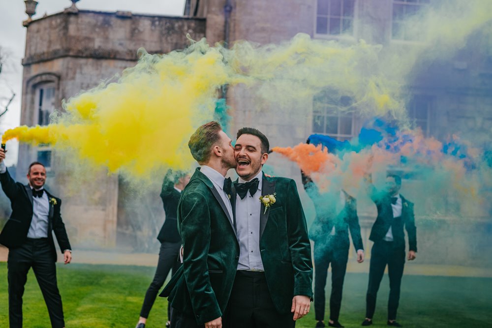 Two grooms kiss on the cheek whilst surrounded by rainbow coloured smoke outside their luxe gay wedding venue