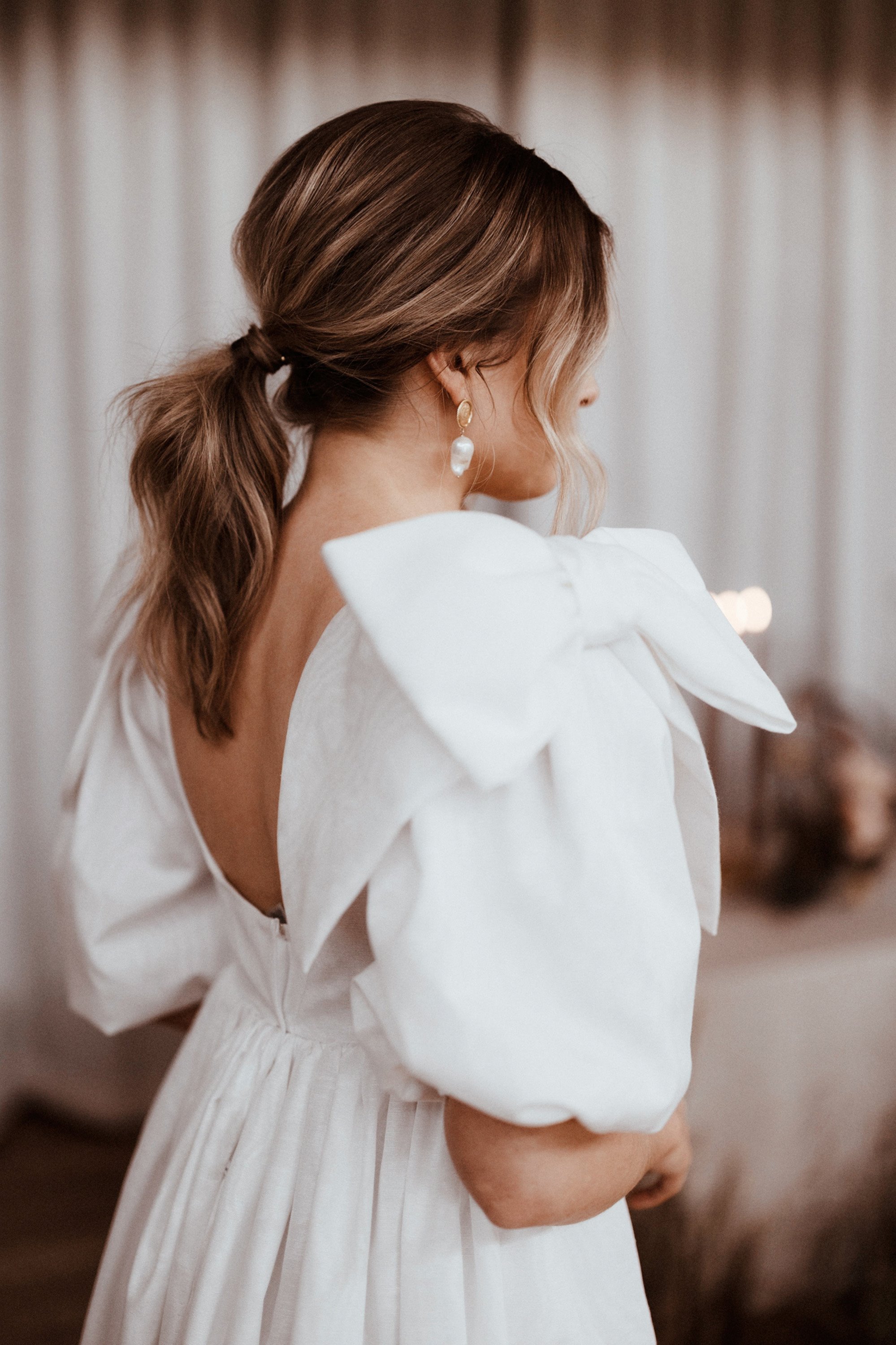 Bride looking like girl with a pearl earring in backless wedding dress with oversized bow no shoulders with modern ponytail hair up