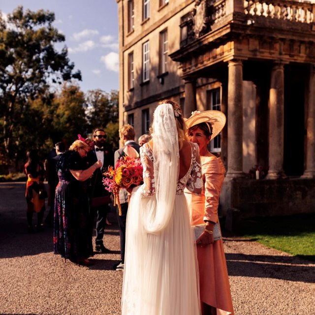 elegant boho bride with long veil standing with guests in the sunshine outside beautiful manor house wedding venue 