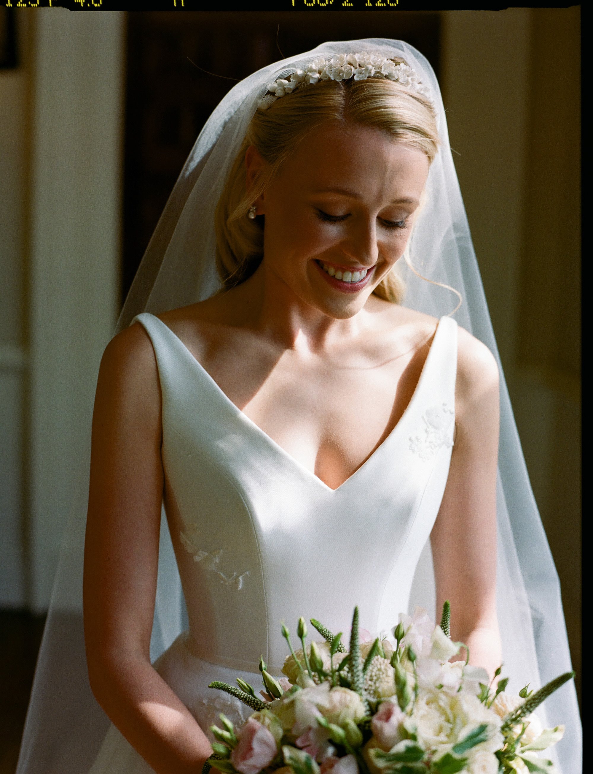 Demure and elegant bride in chic simple satin wedding gown, pearl tiara and veil, pearl necklace and pearl earrings smiles down at her bouquet of white, pale pink and green flowers