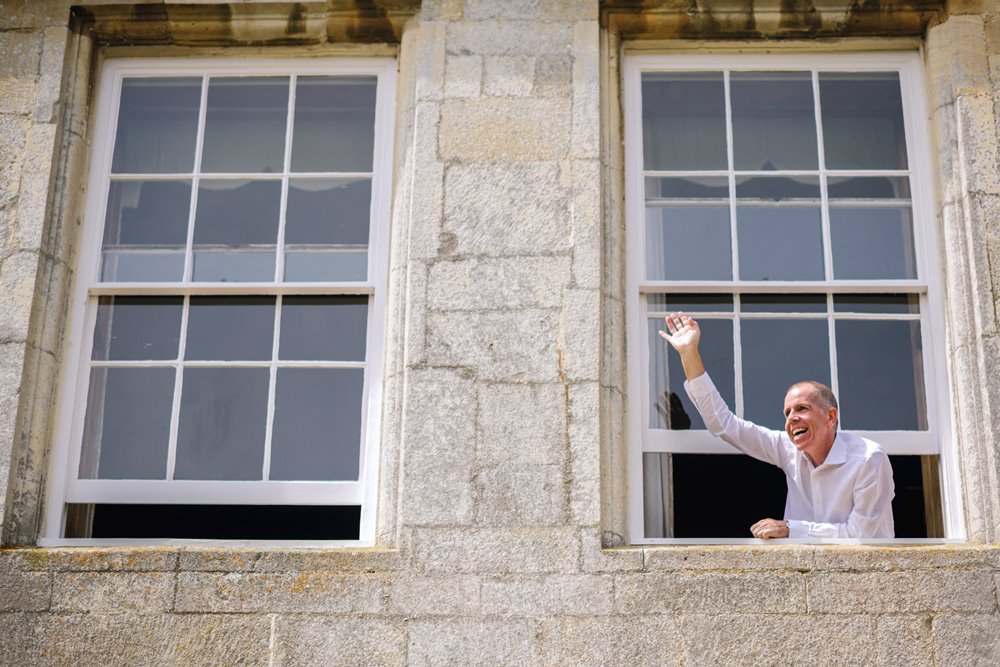 Groom waving out window of mansion house as guests arrive