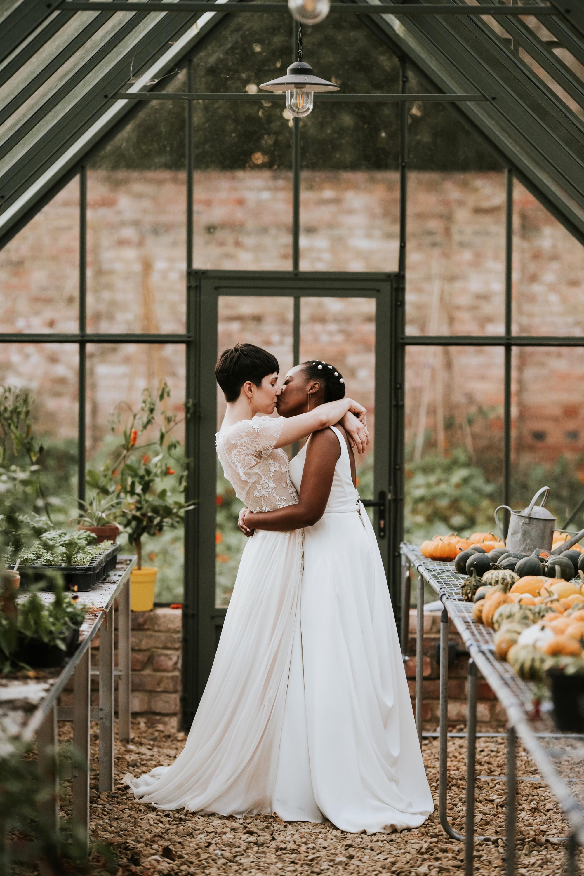 two beautiful brides embracing in a greenhouse of a walled garden, with pumpkins and plants all around them