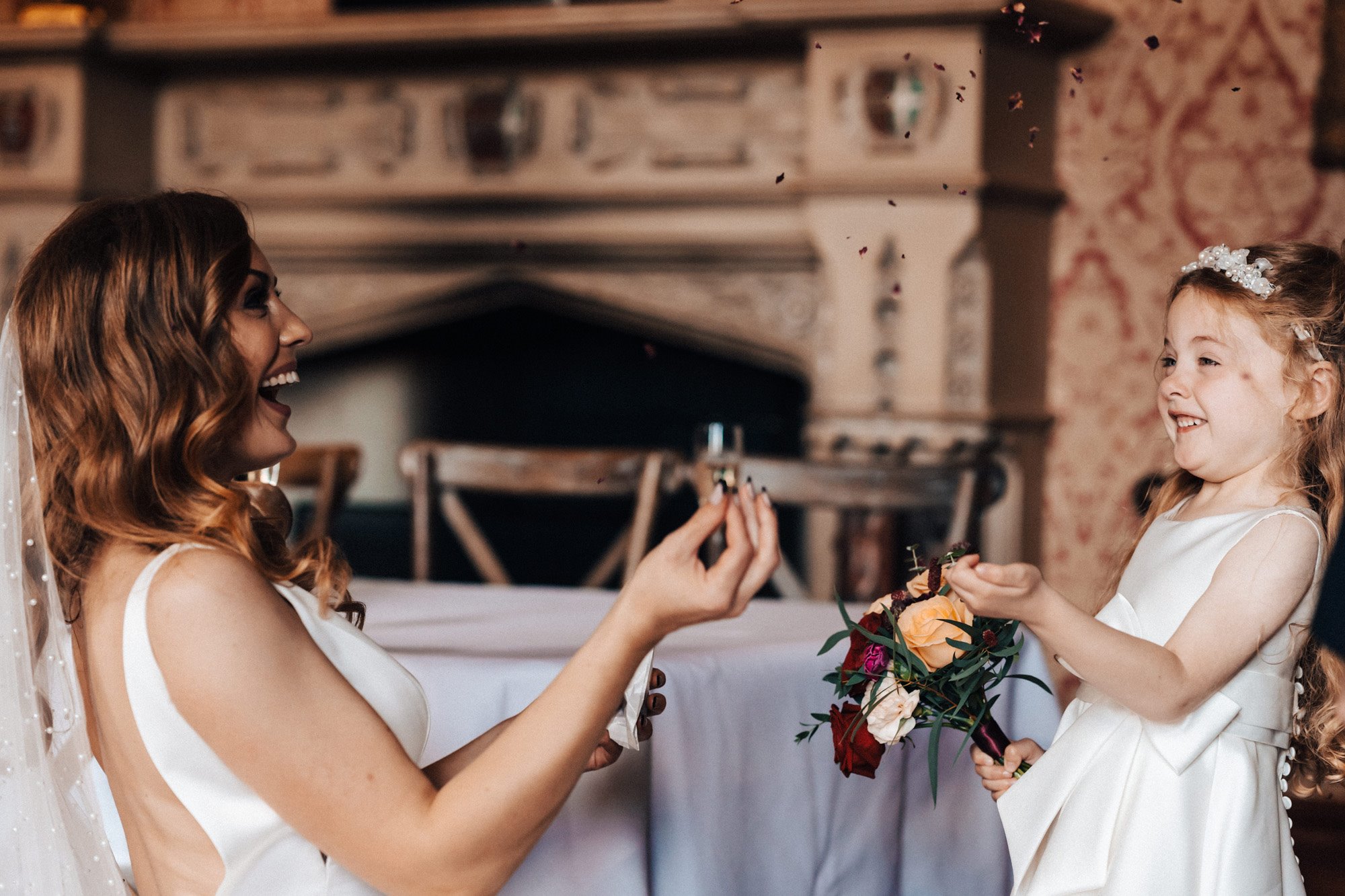 Beautiful bride in pearl veil and little bridesmaid daughter in pearl tiara smile and laugh in historic house with huge fireplace