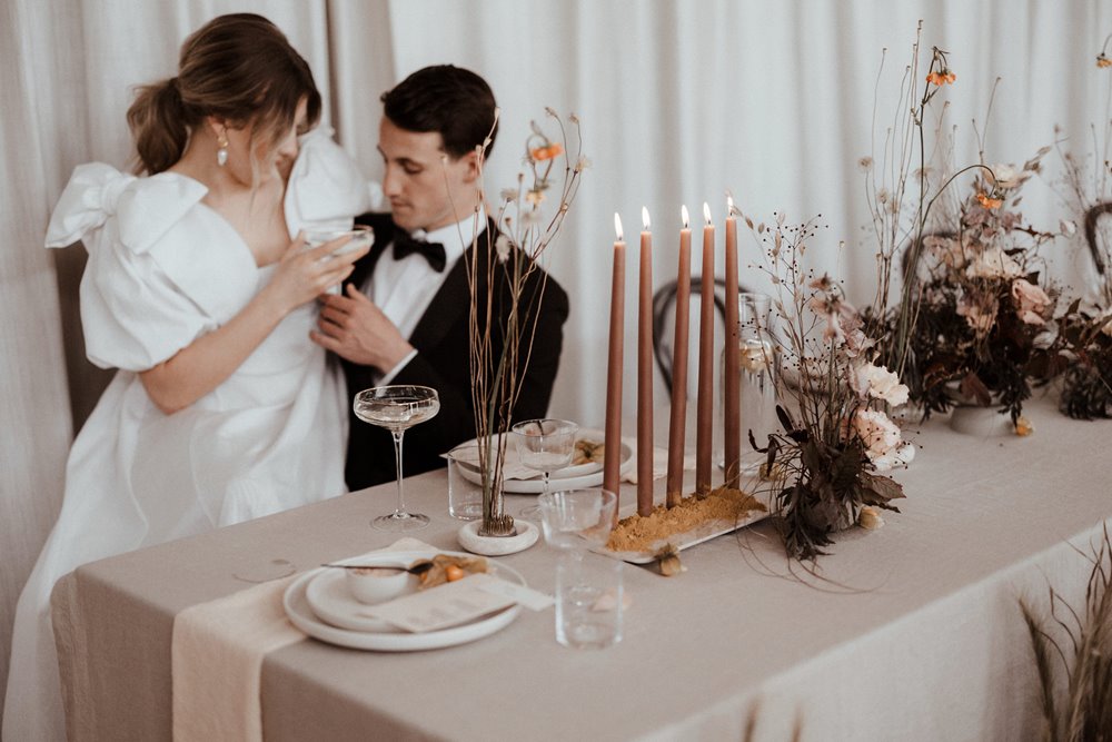 Bride and groom sit at natural wedding table styled with eco details wearing luxe oversized bow wedding dress and tux