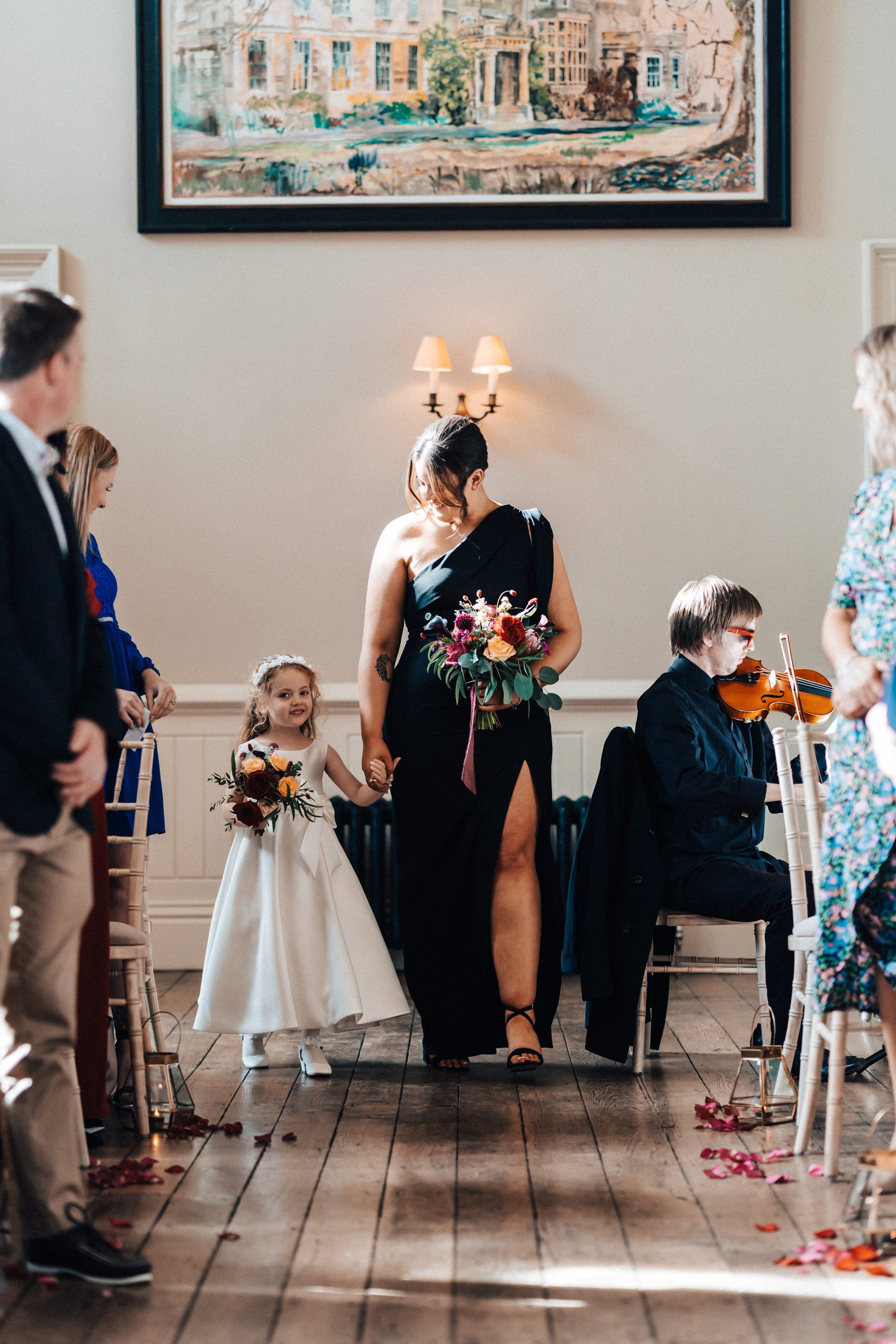 bridemaid wearing black dress walks down aisle holding flower girls hand in an Autumn wedding ceremony at a country house in gloucestershire