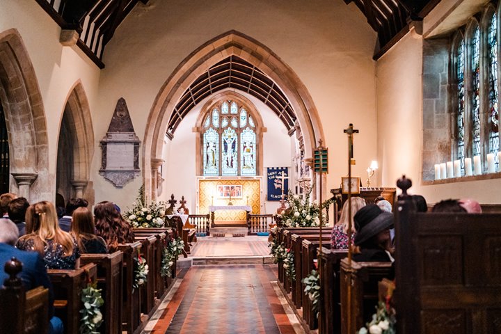 England's prettiest church at a wedding ceremony in Elmore, golden light lights up the stained glass windows as guests sit waiting for the bride and groom to arrive