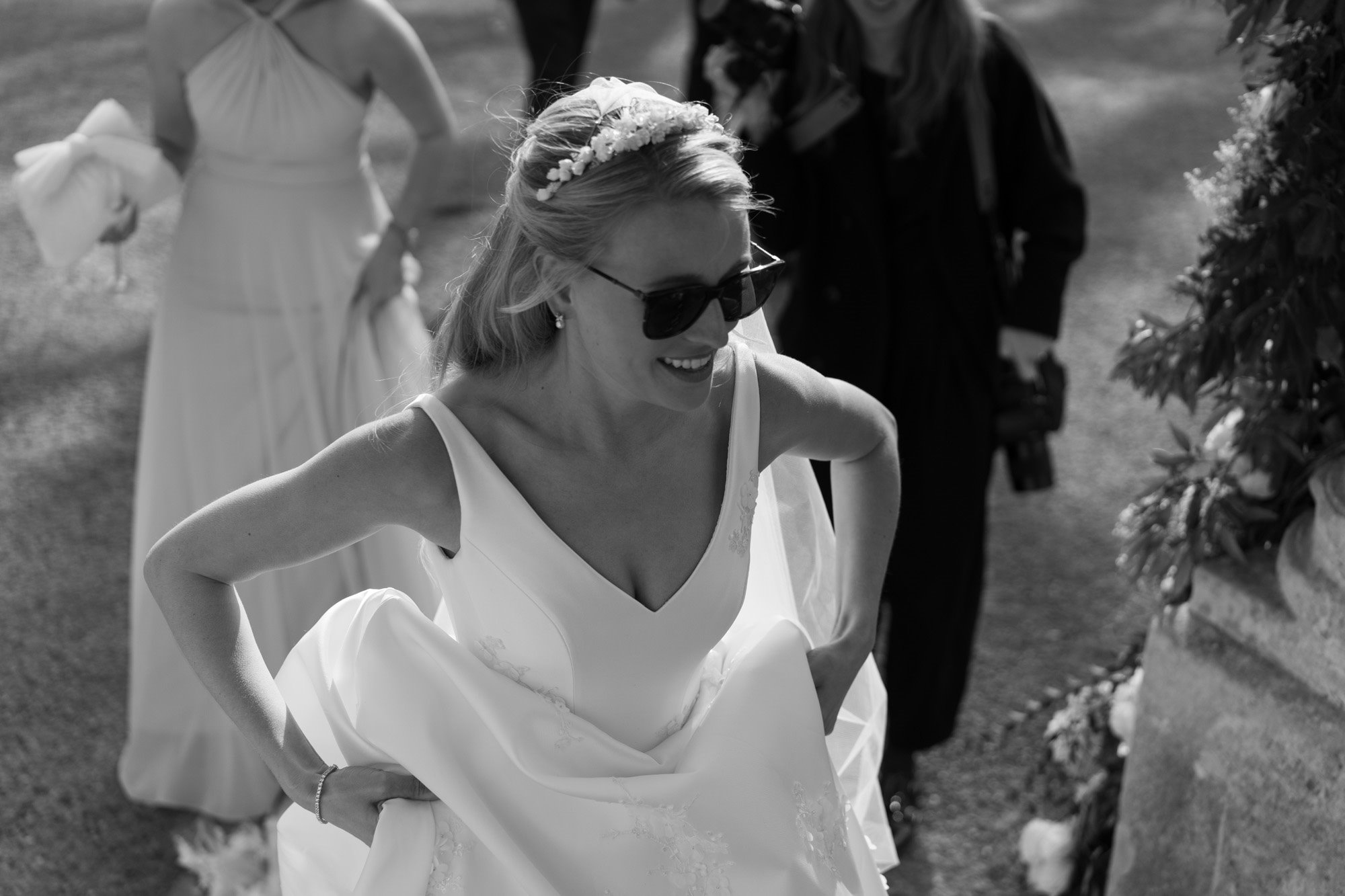 Cool modern bride in Ray Ban sunglasses, pearl bridal tiara, pearl necklace and pearl earrings lifts up the skirt of her princess wedding dress to walk up the steps of stately home wedding venue