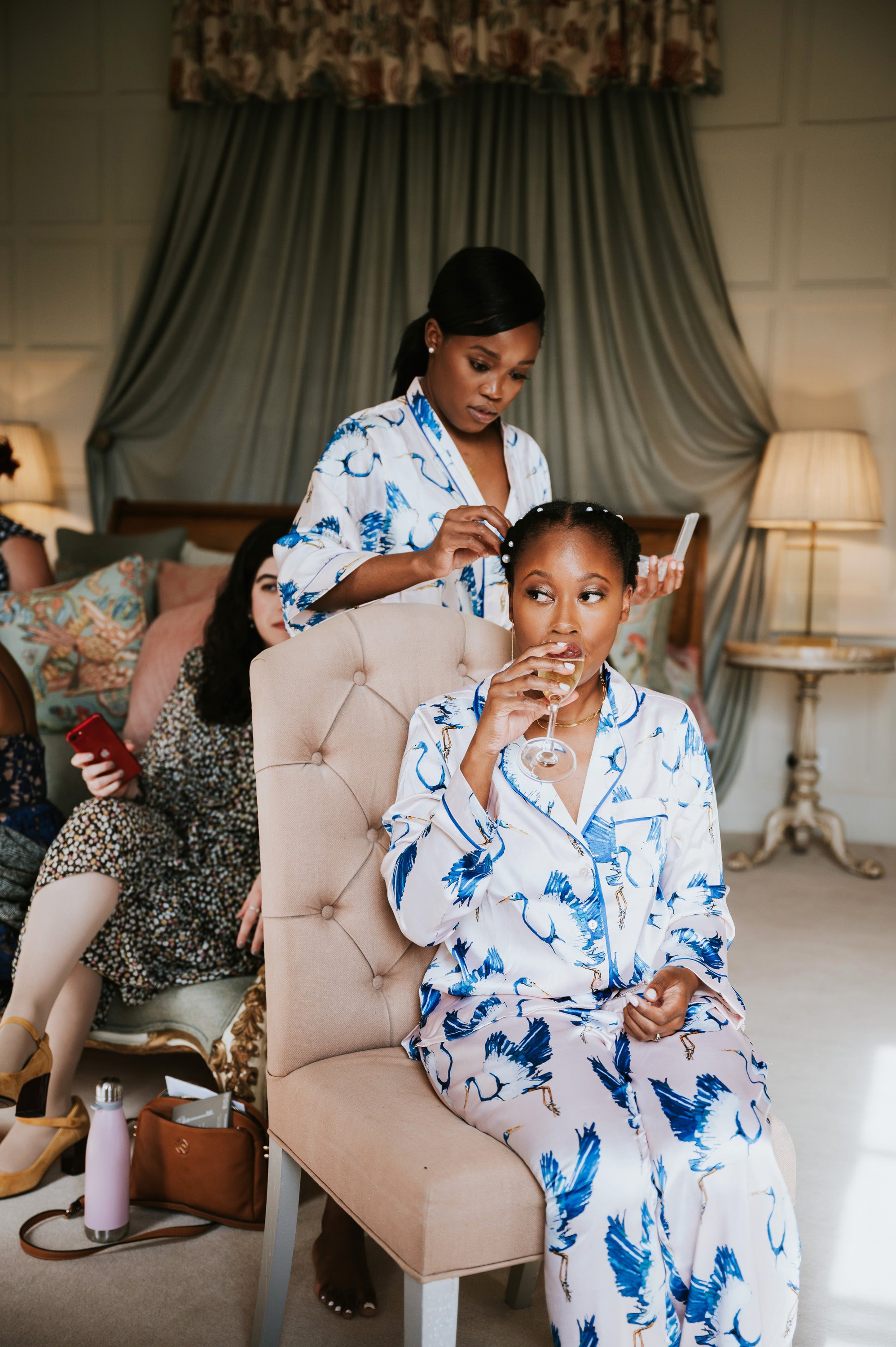 Bride with afro hair sits sipping champagne in silk pyjamas as sister pins pearls in her hair in beautiful luxury stately home bedroom