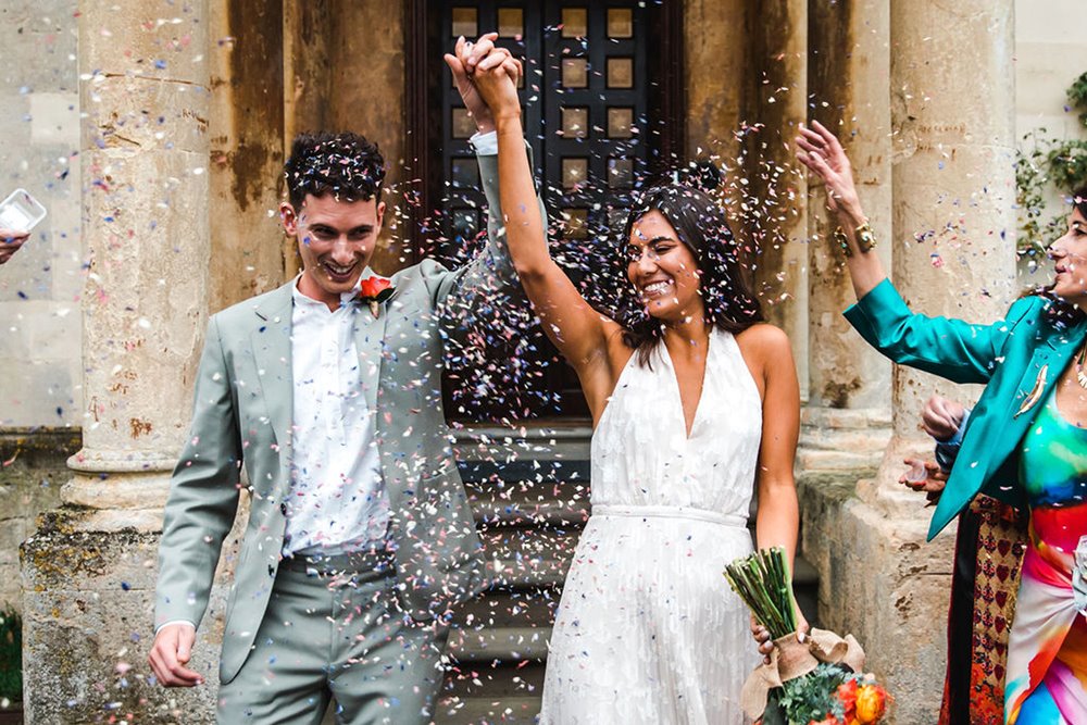 Just married couple smile and hold hands in the air whilst confetti is thrown at micro wedding ceremony at elmore court