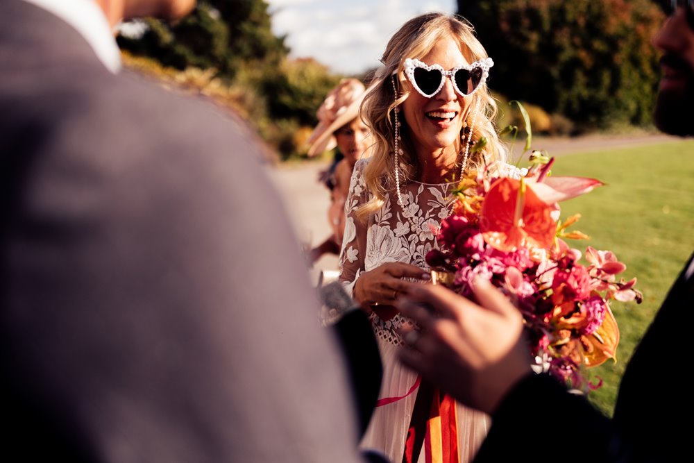 cool bride in heart shaped sunglasses and bright wedding flowers in sunny drinks reception outside elmore court