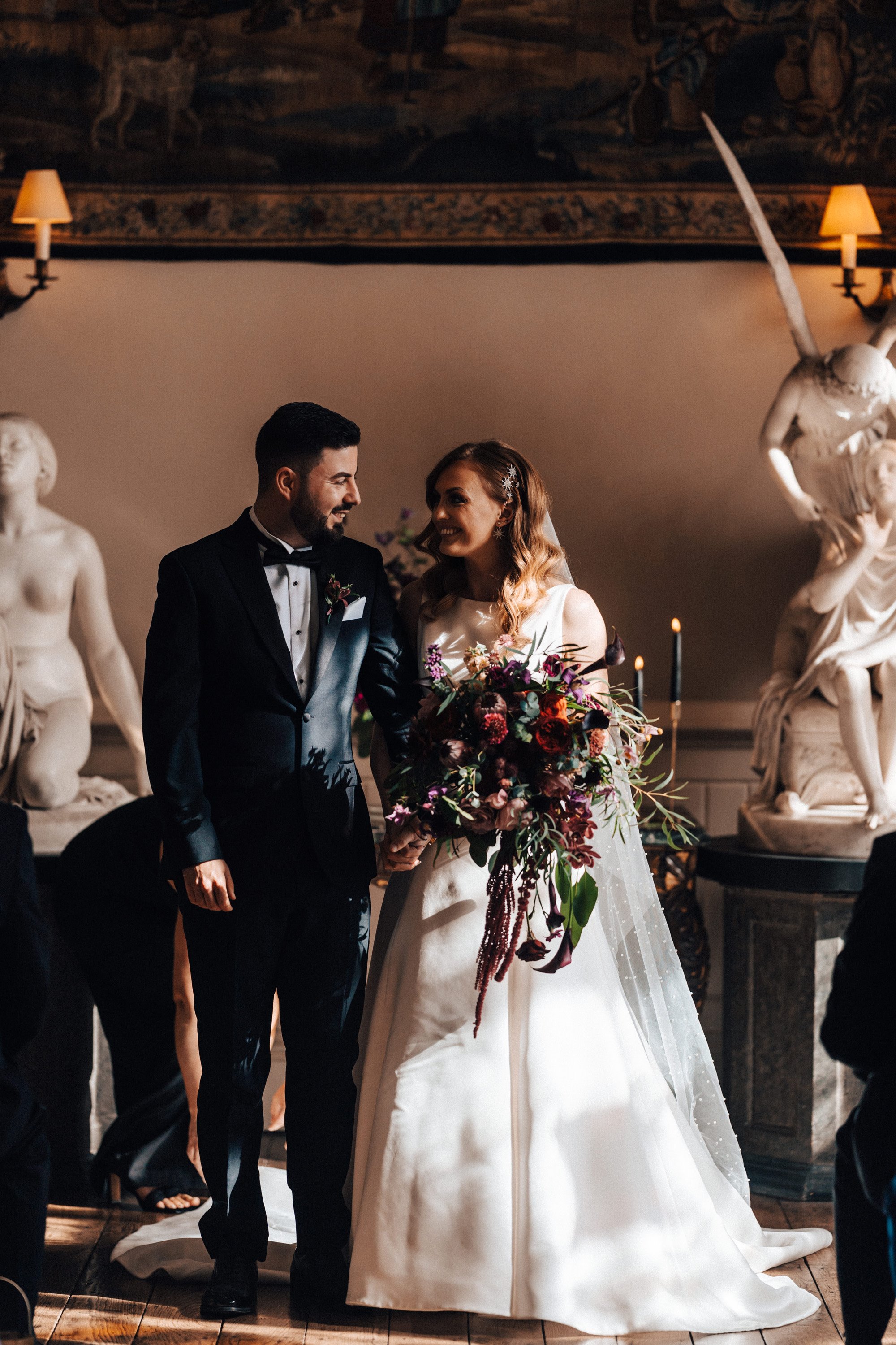 Bride and groom grin happily at each other at the end of a beautiful aisle with angel statues in a historic mansion house in the english countryside