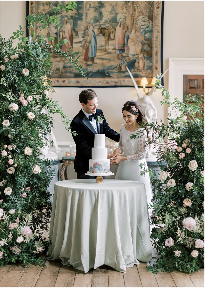 Luxe bride and groom cut rainbow cake surrounded by greenery flower arches in stately home wedding venue in the UK