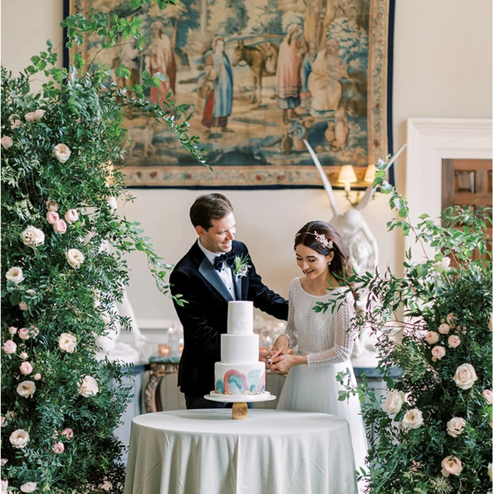 Luxe bride and groom cut rainbow cake surrounded by greenery flower arches in stately home wedding venue in the UK