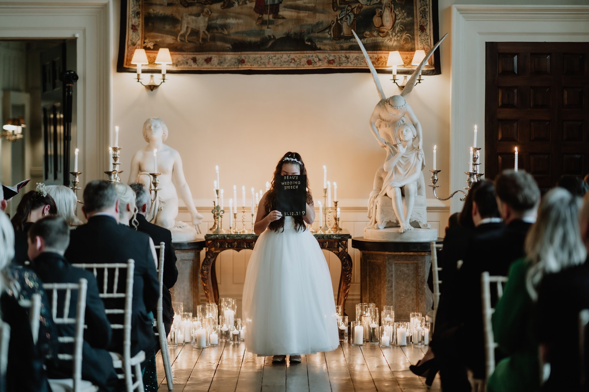 Flower girl posing at the end of the aisle with a stunning display of candles and sculptures behind her