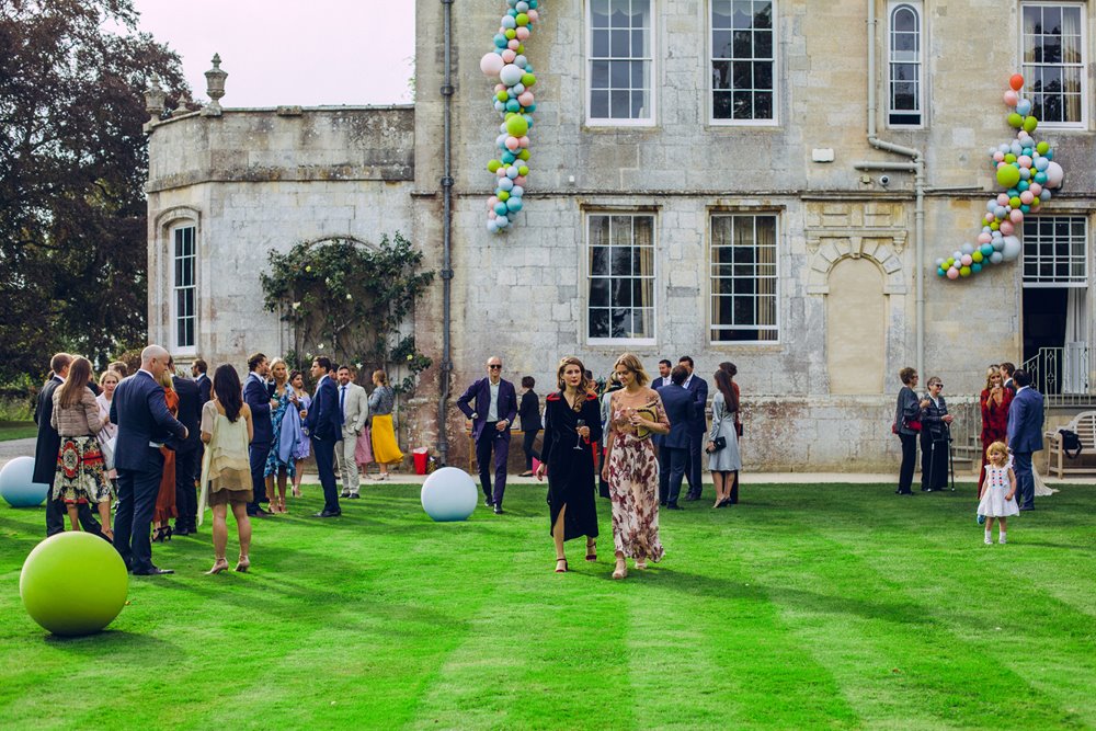 Guests at a big wedding chat on a bright green lawn in front of huge stately home wedding venue in the cotswolds