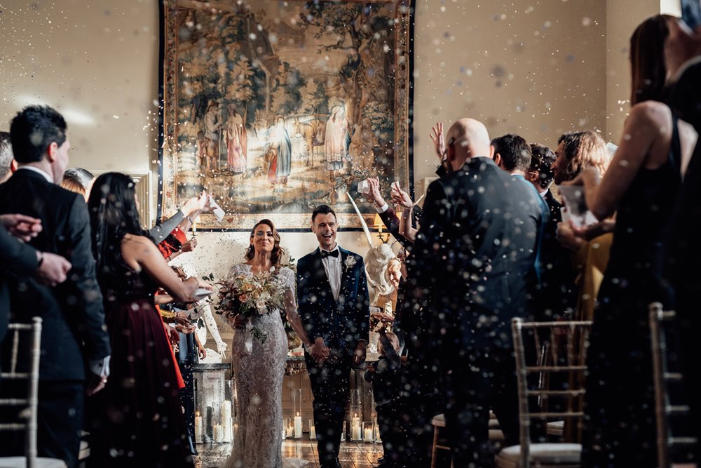 Beautiful couple walk down the aisle in luxury mansion wedding venue with guests throwing confetti on either side