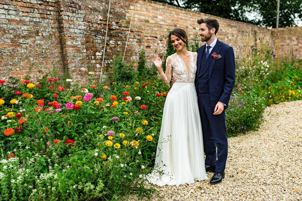 Elegant bride in glamourous french wedding dress stands with her husband next to the flowers in the walled garden at elmore court on their wedding day