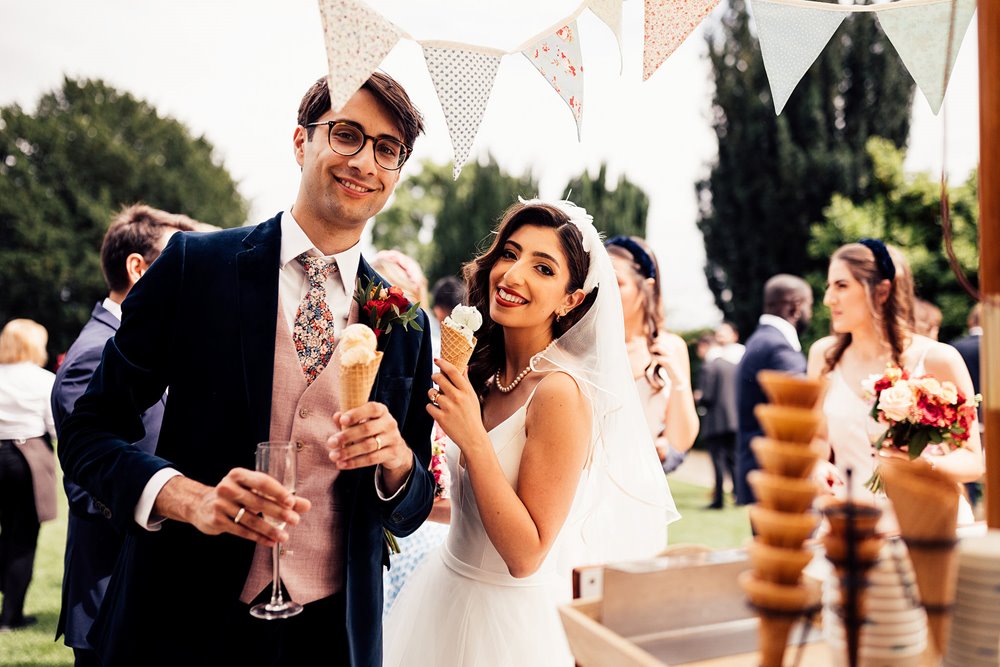 Gorgeous bride and groom pose with ice cream cones under their bunting decorated vintage ice cream stand at their garden wedding in the cotswolds