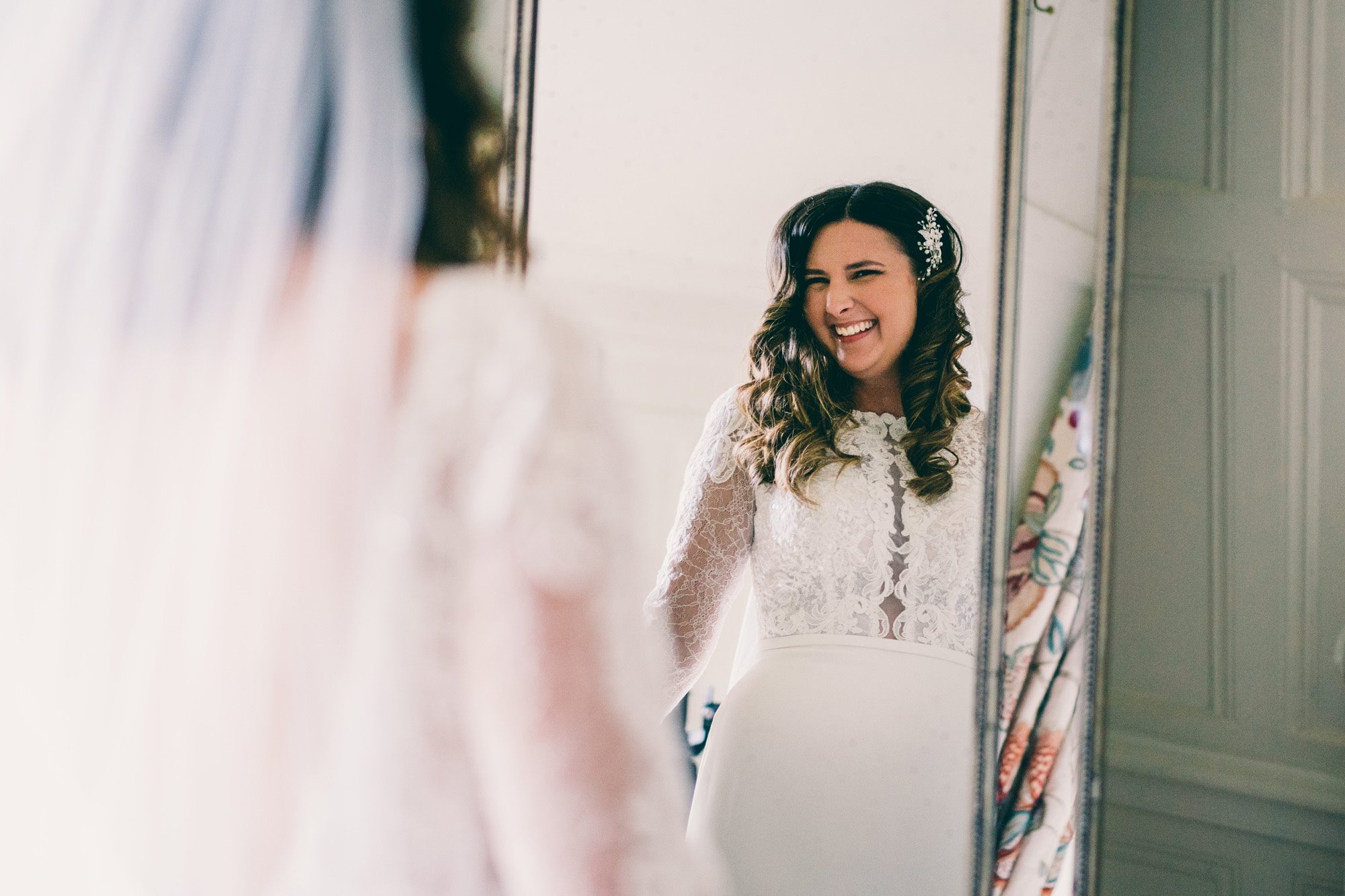 Bride in lace dress smiles in mirror at brunette curly hair with pearl hair clip 