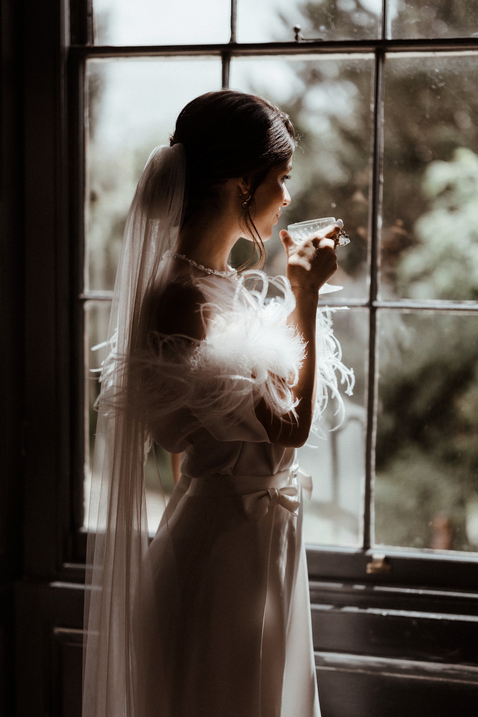 Vogue wedding inspiration bride with chignon and long veil in feathers looks out window of stately home holding crystal glass in this fine art modern wedding editorial