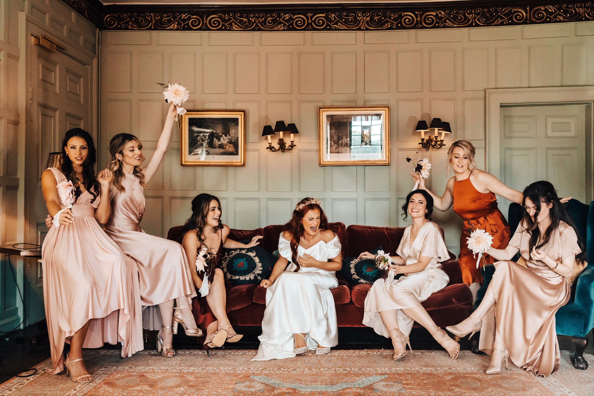 Fun bridal party photo of bridesmaids in pink silk dresses holding up bouquets and bride laughing sitting on red velvet sofa in a stately home wedding venue