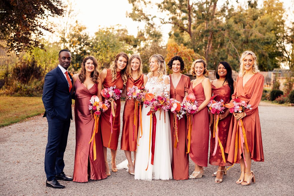 brides man and bridesmaids in burnt orange copper dresses stand with boho bride with bright bouquets outside stately home wedding venue in the cotswolds countryside 